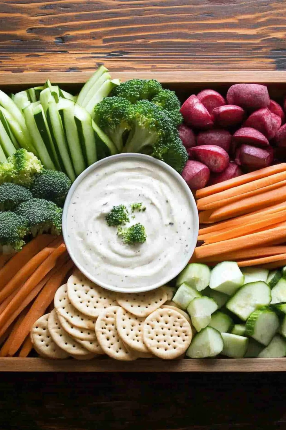 Eine Holzkiste mit frischem Gemüse wie Karotten, Gurken, Sellerie, Brokkoli und Rote Beete, zusammen mit Crackern und einer Schale Dip.