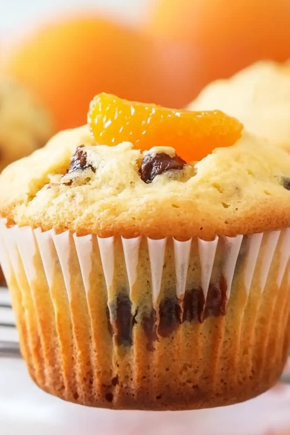 Nahaufnahme eines fluffigen Orangenmuffins mit Schokostückchen, garniert mit einem Stück Mandarine und umgeben von weiteren Muffins im Hintergrund.