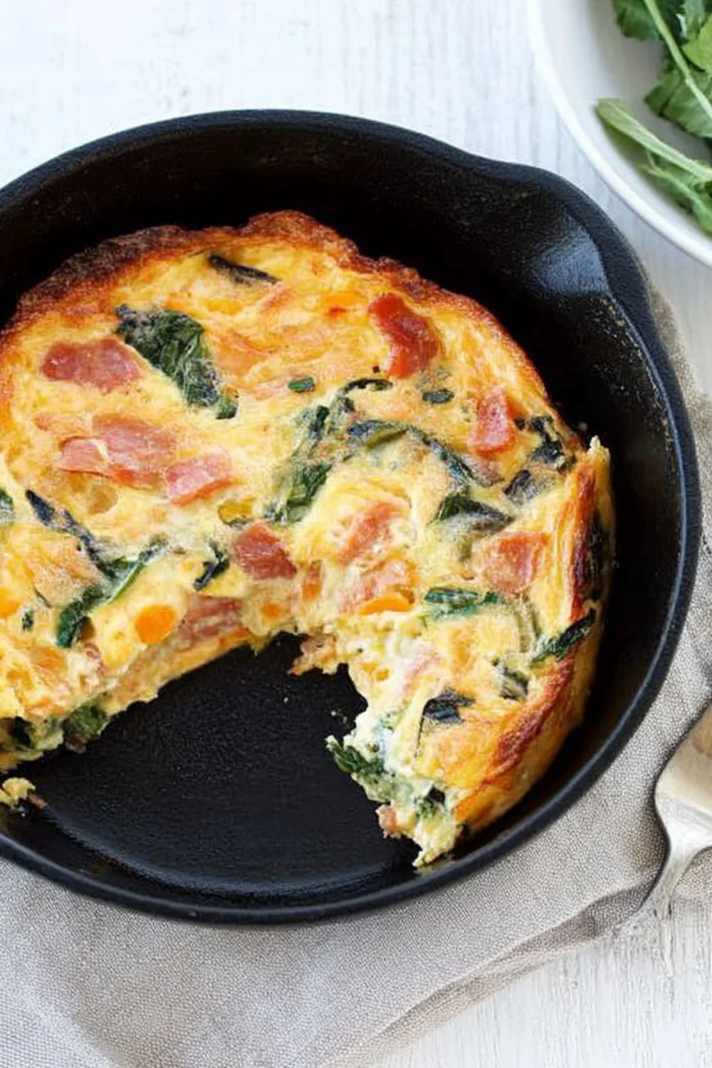 Ein leckerer Gemüse-Bacon-Auflauf in einer schwarzen Gusseisenpfanne, mit einer Portion herausgeschnitten, die eine Mischung aus Spinat und gebratenem Bacon zeigt.