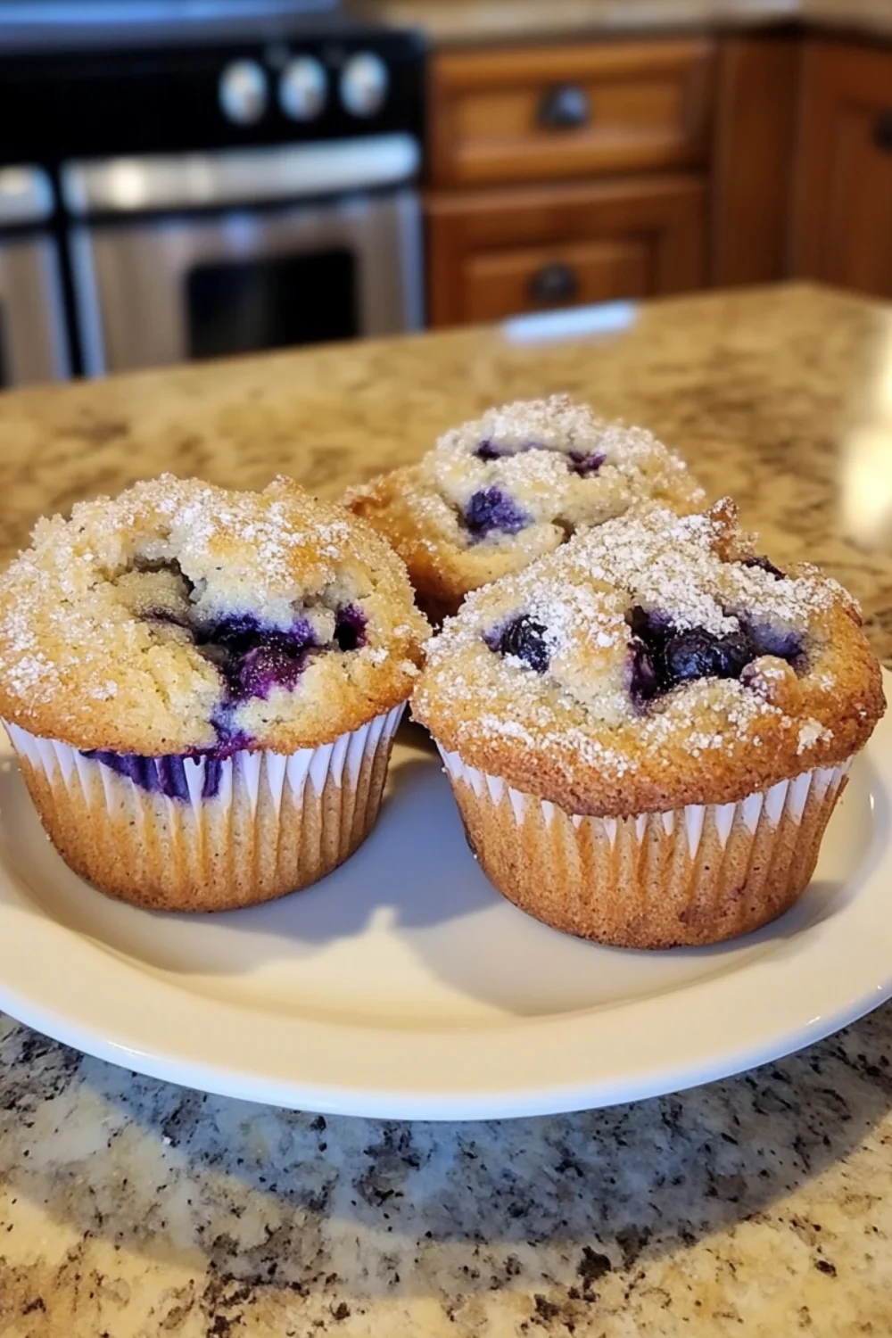 Drei Blaubeermuffins in weißen Papierförmchen auf einem weißen Teller.