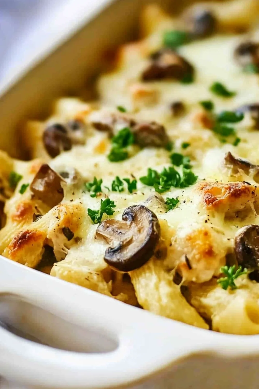 Eine Nahaufnahme eines köstlichen, gebackenen Hähnchen-Mushroom-Pasta-Auflaufs, garniert mit frischer Petersilie, in einer weißen Auflaufform.
